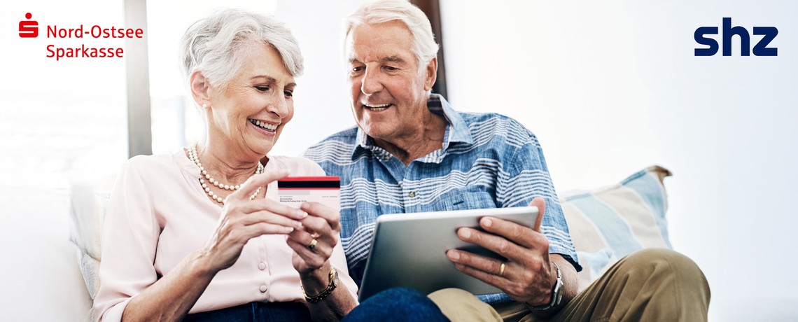 Ein älteres Paar zeigt sich gegenseitig eine Karte der Nord-Ostsee Sparkasse und ein Tablet mit der digitalen Tageszeitung des shz